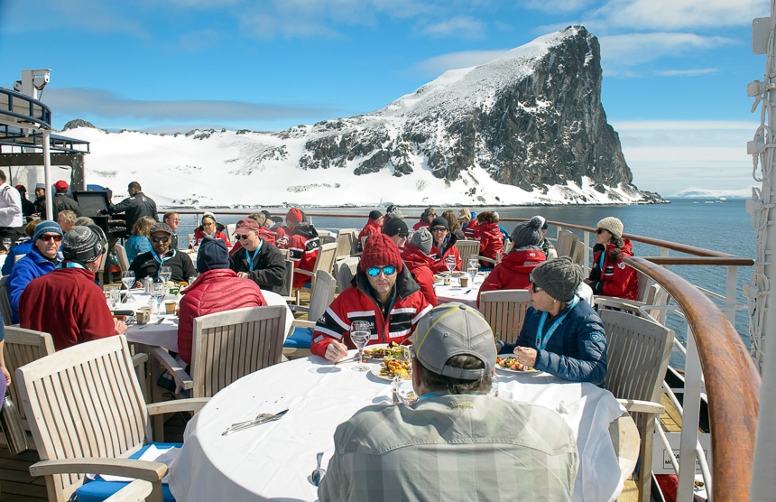 On a sunny blue sky day in Antarctica guests sit on the top deck of the ship and enjoy an outdoor BBQ lunch in front of a snow covered jagged rock formation.