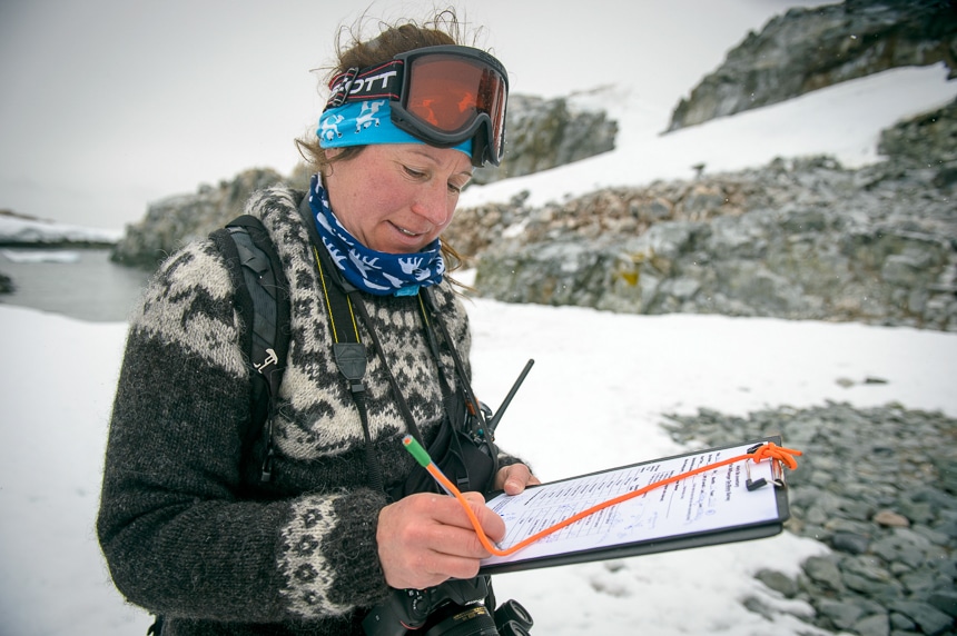 On shore in Antarctica a naturalist guide from Polar Latitudes cruise line wears snow goggles and looks down as she writes on a clipboard