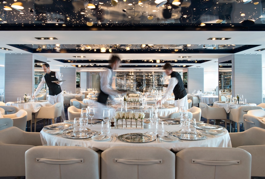 An elegant dining room aboard a luxury Antarctica cruise line filled with white tables clothes, white roses and cream leather chairs sit empty as 3 crew members set the table ware.