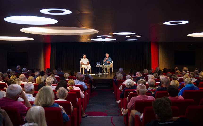 Guests sit in theatre style seating facing the stage watching two guest speakers give a lecture aboard a ponant cruise line ship.