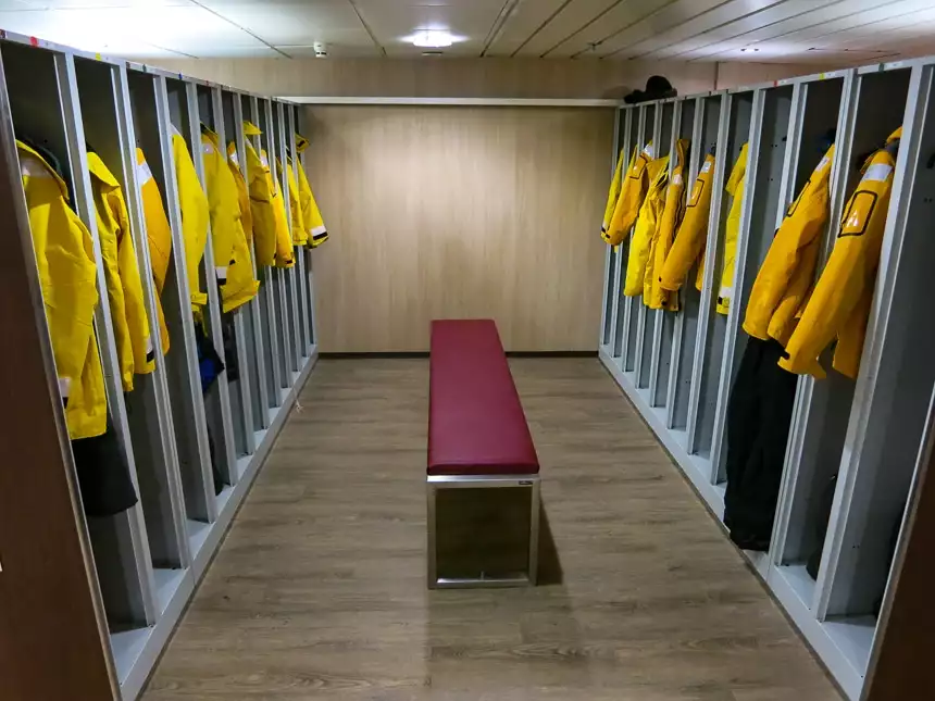 Mud room aboard Quark Antarctica cruise line ship. Yellow parkas are hung up in open long cubbies with a bench seat on the floor.