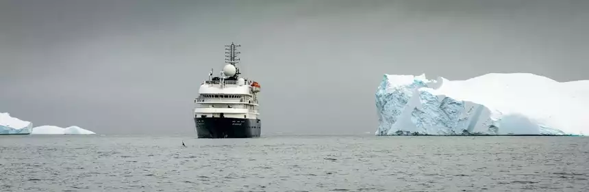 On a grey overcast day in Antarctica a white and blue ship navigates the polar ocean next to a massive white iceberg.