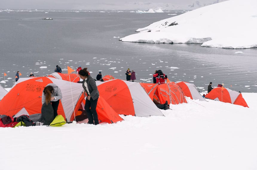 polar travelers set up orange & gray tents atop a snowfield with iceberg-filled waters behind while camping in antarctica.