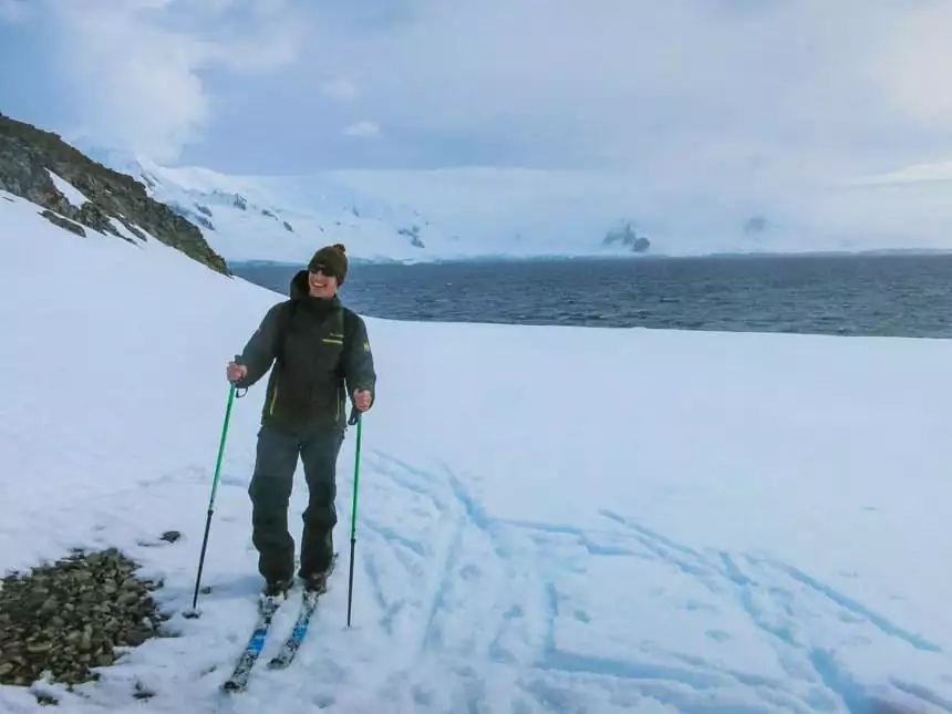 Man in dark green jacket, pants & knit hat stands on nordic skis atop snow with ocean behind, cross country skiing in antarctica.