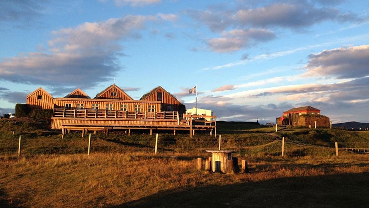 Long wooden building with sod roof, large deck & Iceland flag flying, on rolling grassland with farm equipment, at dusk.