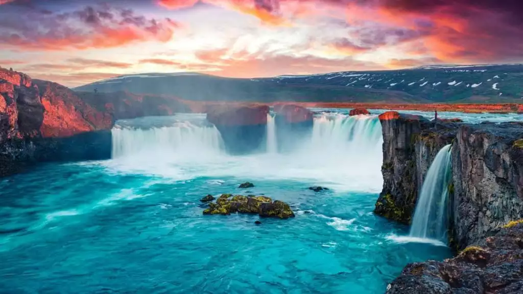 Huge Iceland waterfall sends water down rocky cliffs into a teal lagoon while a traveler stands at the edge at sunset.