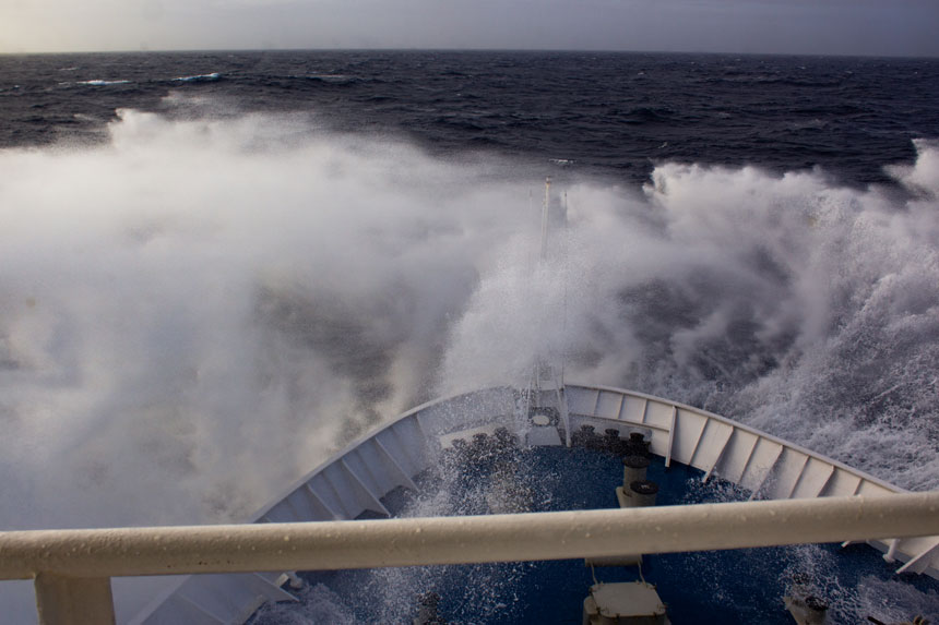Bow of blue & white ship slams down into rough seas while crossing the Drake Passage, one of the places in Antarctica.