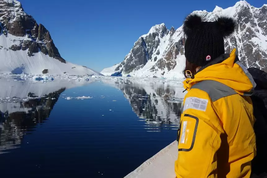 Woman in yellow jacket stands on ship bow looking at the Lemaire Channel a stretch of glassy water with snow-covered, rocky mountains on either side, on a sunny day in Antarctica.