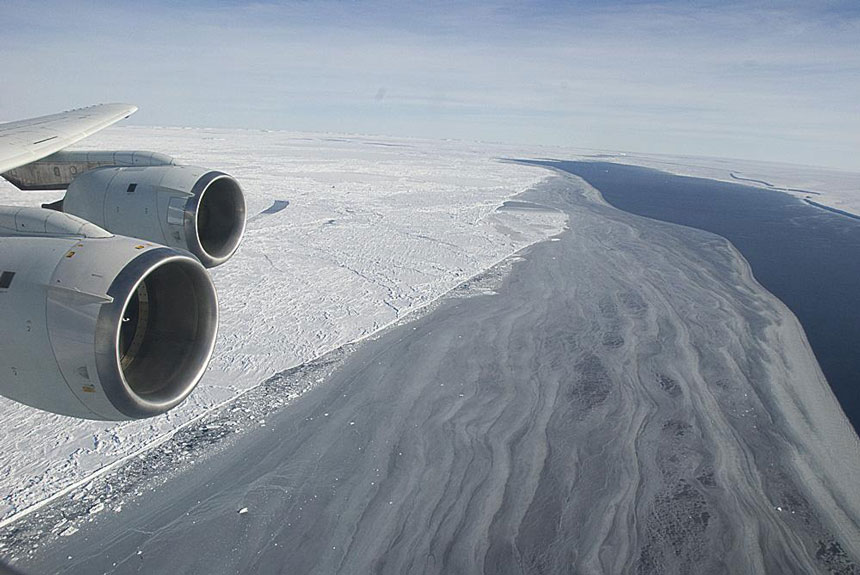 View out a plane window just behind the wing, looking down on the Larsen Ice Shelf, a place in Antarctica, with endless miles of snow tundra & a small inlet of water.