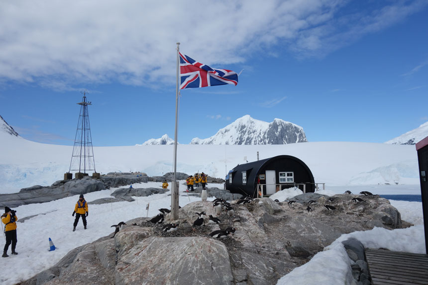 British flag flies outside a dark tube of a building with penguins on rocks in front & people in yellow jackets beside on a sunny day at Port Lockroy in Antarctica.