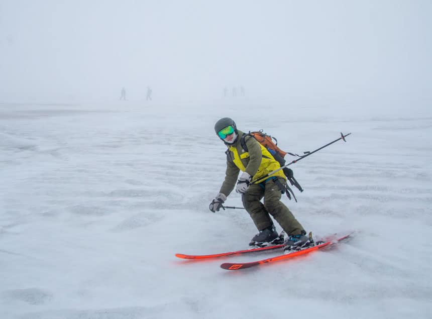 Skiier in green & yellow outerwear, green reflective goggles & olive helmet goes skiing in antarctica down a snowfield on a foggy day with bright orange skis & silver ski poles. 