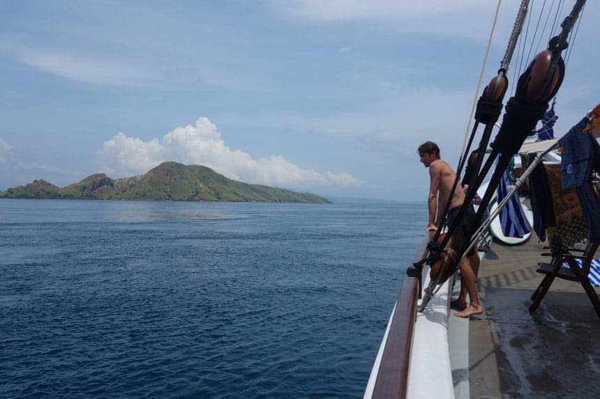 A man leans on the side of a wooden ship in Indonesia with the blue ocean and green island in the background.