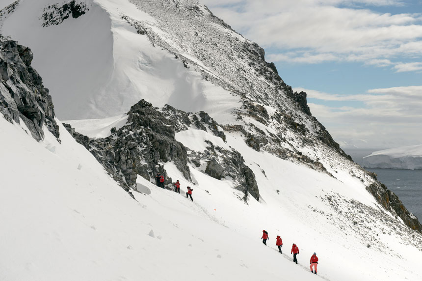 polar travelers in red jackets go trekking in antartica uphill toward a rock band, high above the water, on a sunny day.