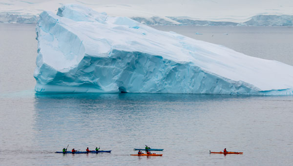 Group of kayakers paddles past an enormous iceberg in calm seas on an Antarctica trip.