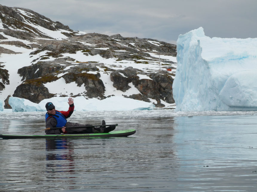 Man lays down & waves from atop a bacl & green stand-up paddleboard while floating in calm water beside an iceberg in antarctica.