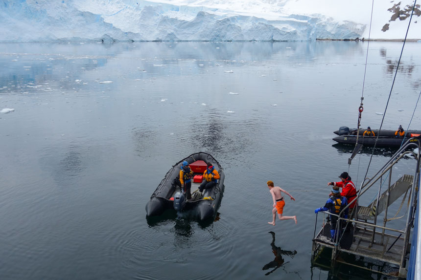 Red-headed man in orange swim trunks jumps off water-level platform into freezing antarctica waters with snow field in background & 2 black Zodiacs with brightly-dressed guides watching.