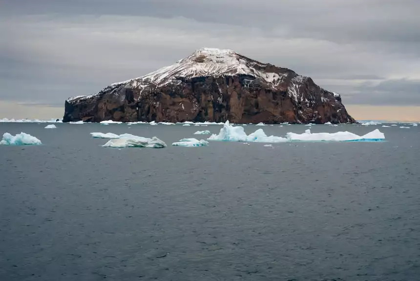 View approaching Paulet Island, a place in Antarctica, with its volcanic crater in the center, brown cliff walls & ocean with small icebergs floating out front.