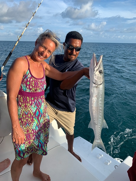 Standing on a boat a woman wearing a patterned dress and the sailboat captain hold a fish that they caught in Belize.