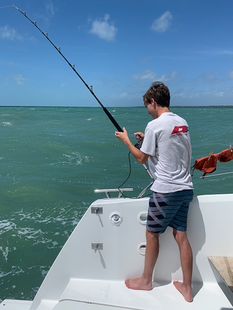 A boy stands on the back of a white catamaran holding a fishing pole for a fishing activity during a Belize charter vacation.