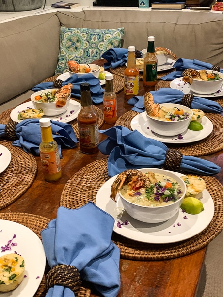 A wooden table with 8 dinner plates of lobster soup, set with 5 hot sauce bottles and bright blue napkins.