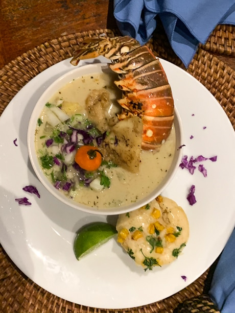 A bowl of soup topped with vegetables and a whole fresh lobster sits on a white plate with a corn patty and line wedge.