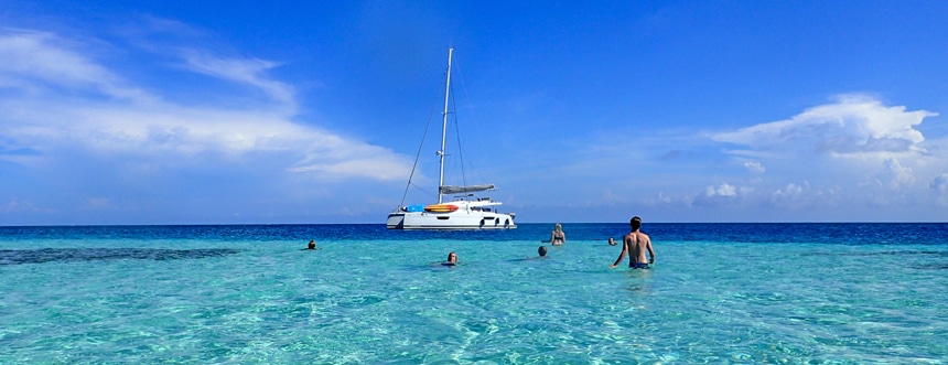 On a bright sunny blue sky day a group of people swim around a white Belize sailboat that floats on the teal blue ocean horizon.