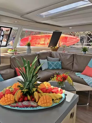 Indoor lounge area aboard Belize catamaran. Upholstered bench seating around a coffee table and a kitchen islands with a plate fresh fruit.
