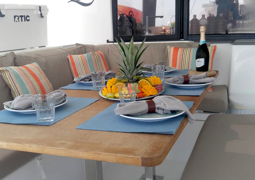Outdoor marine dining table and seating area aboard a Belize catamaran. Table is set for 5 people with a plater of fresh fruit in the middle.