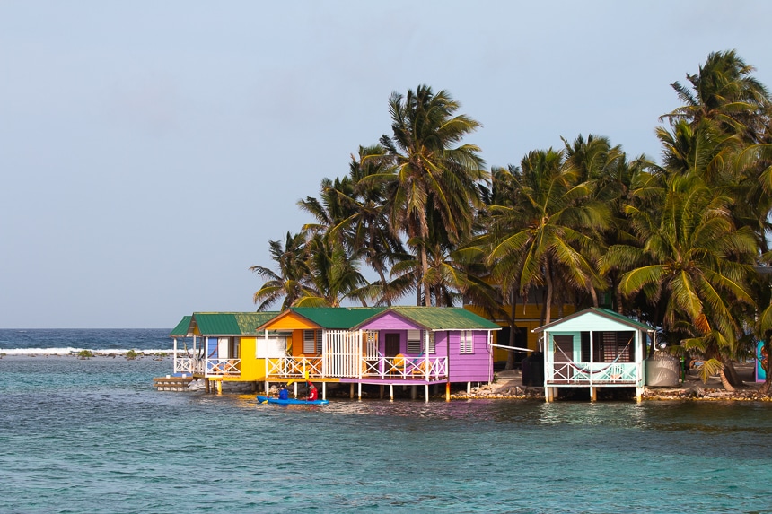 Brightly pained houses on stilts hover over the ocean water on the shoreline of a palm tree filled island called tobacco Caye in Belize.