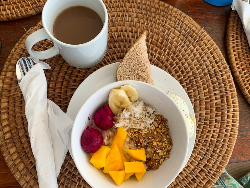 Breakfast of granola, coconut shavings, and brightly colored fresh fruit over yogurt in a white bowl with a slide of bread and a cup of coffee.