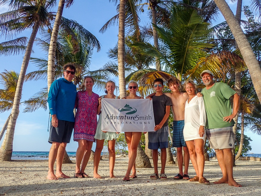 Two families pose together on a sandy palm tree filled island in Belize and hold the AdventureSmith Explorations Flag.