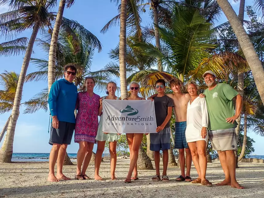 Two families pose together on a sandy palm tree filled island in Belize and hold the AdventureSmith Explorations Flag.