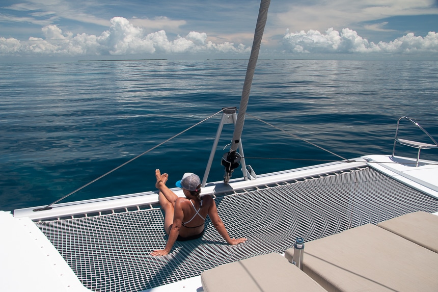 A woman in a bathing suit sits on the netted trampoline at the back of a Belize catamaran looking out over the dark blue ocean.