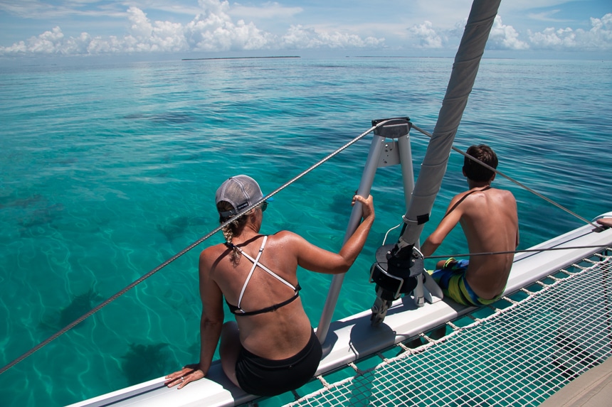 A male and female wearing bathing suits sit on the edge of a Belize catamaran and look out over the teal ocean water horizon of Belize.