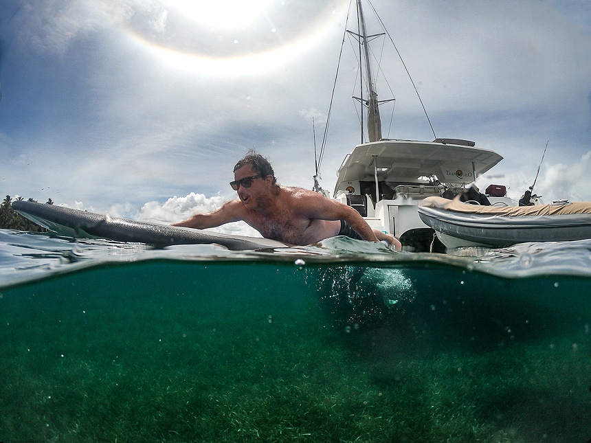 A above and below water photo showing a man paddling a surfboard away from a white catamaran floating in the ocean of Belize.