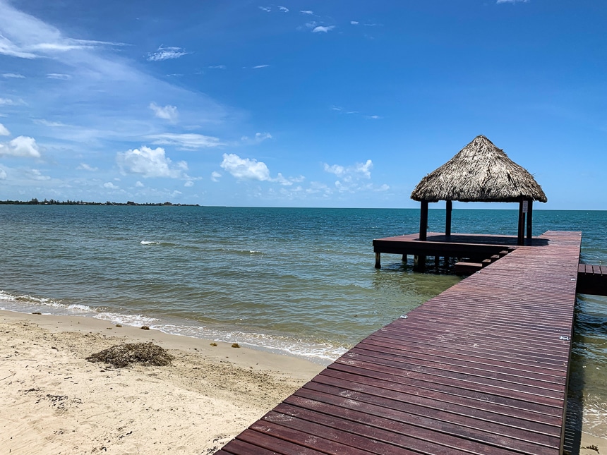 A dark red wooden dock extends into the blue ocean from the sandy shore in Belize, with a thatch roof cabana at the end.
