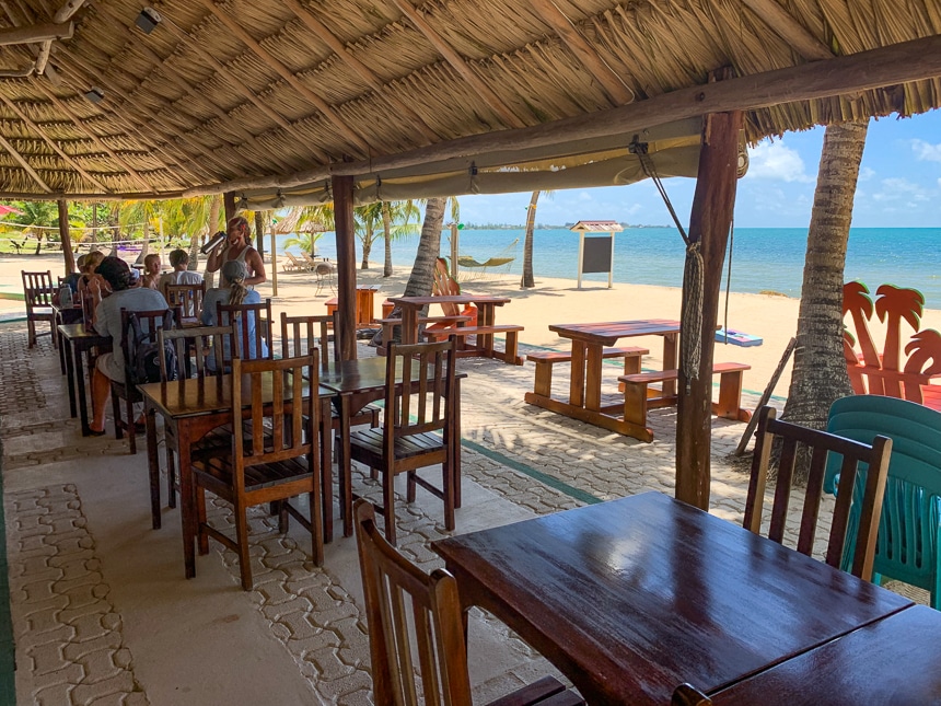 An open air and ocean front restaurant with a thatch roof and wooden tables and chairs shown on bright sunny day on an island in Belize.