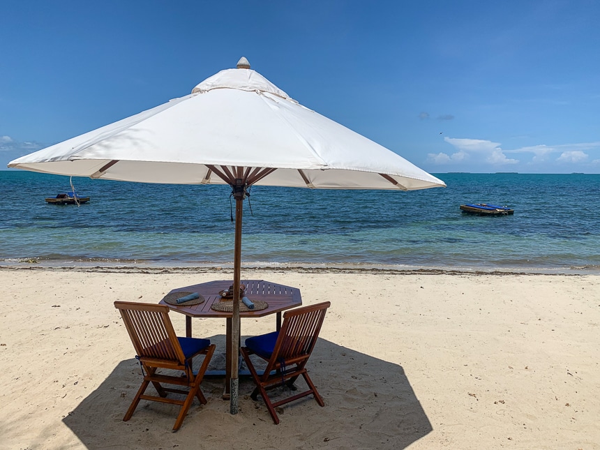 In Belize a small table for two is under a large white umbrella set on the beach with views of the blue teal ocean and sky.