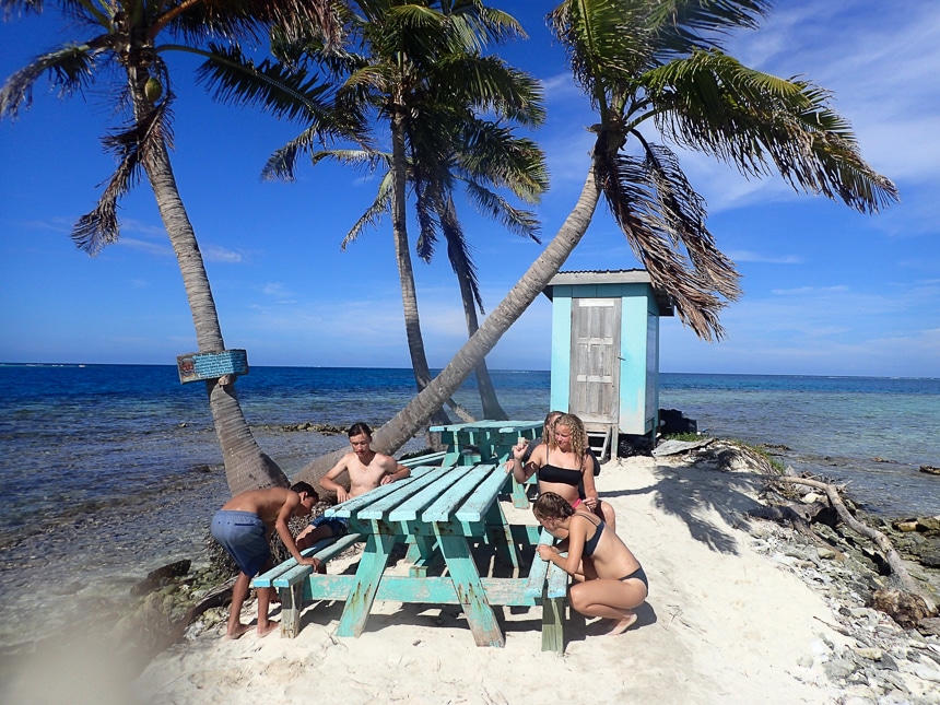 Children in bathing suits sits around teal wooden picnic tables on a tiny sandy islands of palm trees during a Belize vacation.
