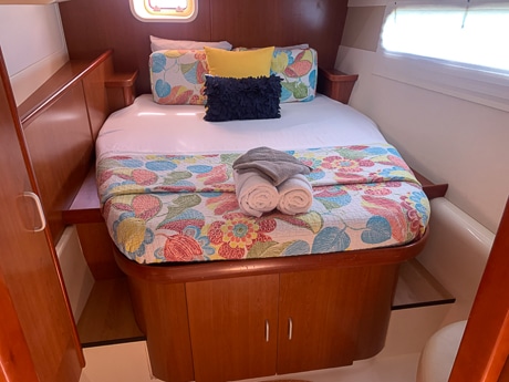 Inside a cabin aboard a Belize sailboat. One long window, a wooden bedframe with cabinets sits underneath a mattress wrapped in colorful blankets.
