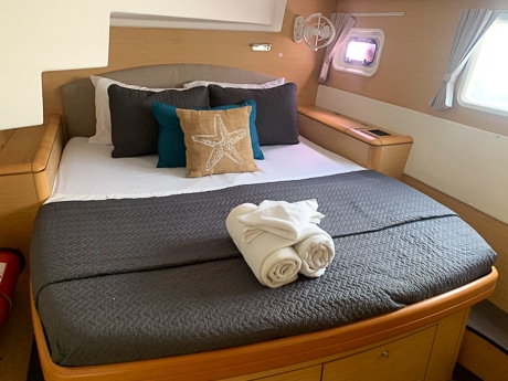 A windowed cabin aboard a Belize sailboat. Includes a wooden bedframe with cabinets underneath a mattress wrapped in a blue blanket and rolled towels on the end.