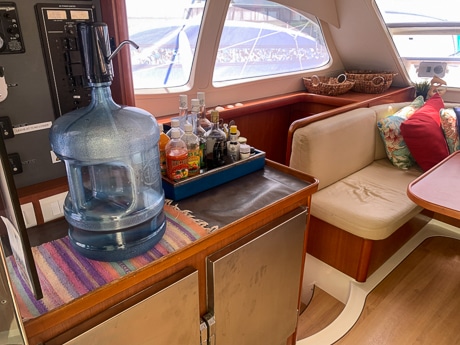 The indoor salon dining area aboard a Belize sailboat. Upholstered bench seating around a table with a large waters jug and tray of condiments on the counter.