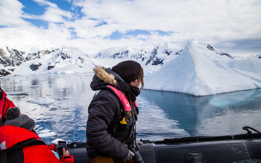 Female traveler wearing a black jacket holds camera and sits inside a zodiac boat cruising through floating Antarctica icebergs.