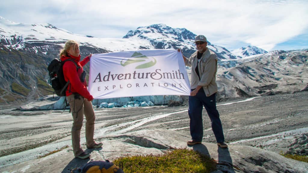 Two travelers holding an AdventureSmith Explorations flag in front of Kenai Fjords glaciers on a Denali vacation