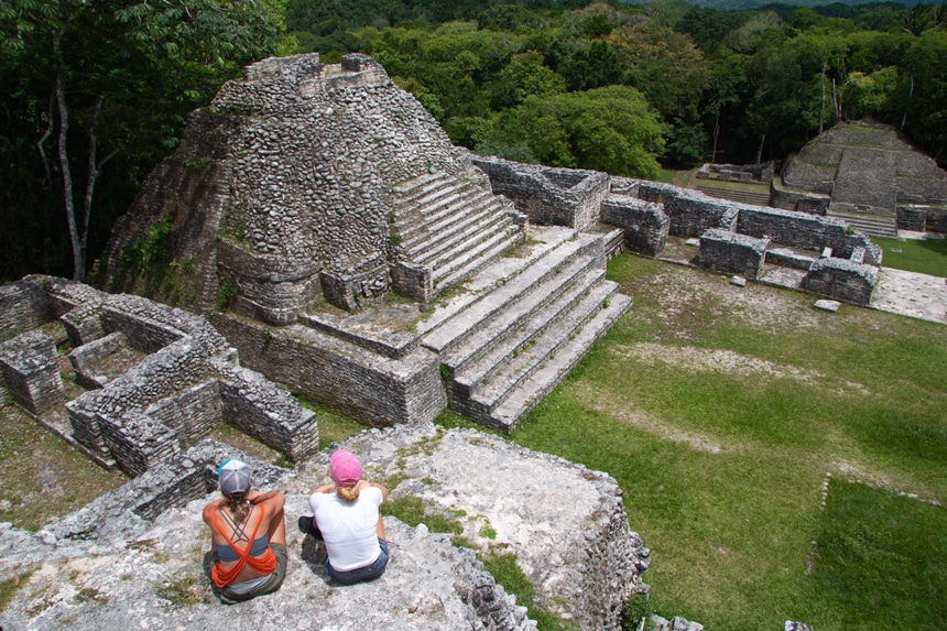 Two female travelers sit and look down over a set of stone Mayan temple ruins in Belize surrounded by green lush jungle.