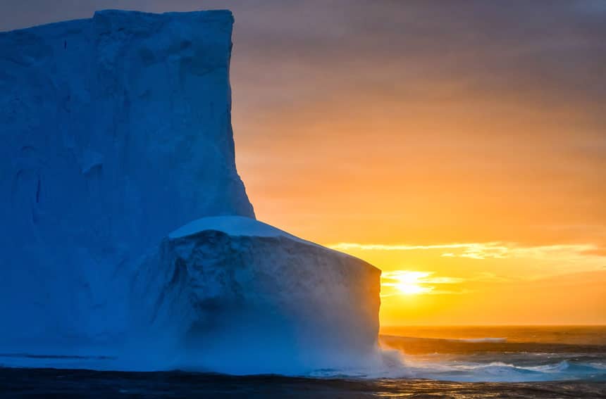 Sun sets behind a very tall blue shelf of ice known as the Ross Ice Shelf, one of the places to visit in Antarctica.