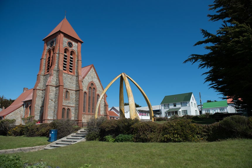Stone church with brick-red trim surrounded by green grass & hedges, white houses with green roofs & an orb-like gold arch, on a sunny day in Port Stanley, Falklands.