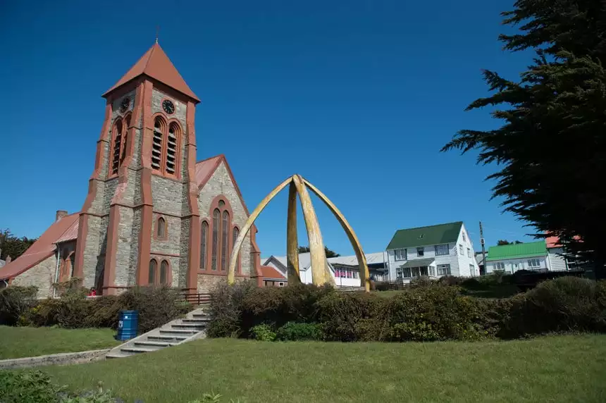 Stone church with brick-red trim surrounded by green grass & hedges, white houses with green roofs & an orb-like gold arch, on a sunny day in Port Stanley, Falklands.