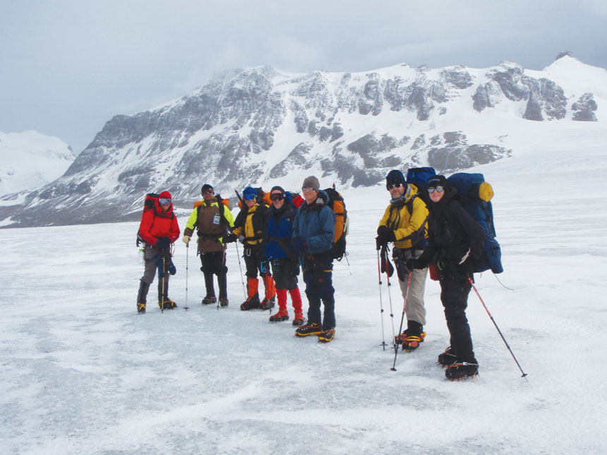 Group of polar travelers with large packs & trekking poles stand atop a snowfield during the Shackleton Crossing on South Georgia Island.