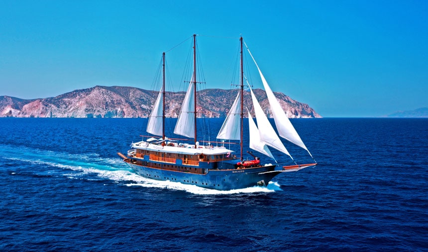 Classic motorsailor with white sails, wooden upper deck & blue hull cruises in open water beside Mediterranean islands.